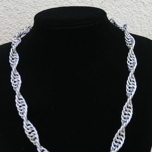Puede incluir: Un collar de cadena de plata con un diseño trenzado. El collar se muestra en un soporte de terciopelo negro.