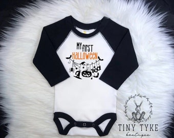 Puede incluir: Un body blanco de manga larga para bebé con mangas negras. El body tiene un gráfico naranja y negro que dice "Mi primer Halloween" con una cadena de decoraciones de Halloween colgando de él. El body tiene un borde negro alrededor del cuello y la parte inferior. El body está sobre una superficie blanca y esponjosa. El texto "Tiny Tyke Boutique" está en la esquina inferior derecha de la imagen.