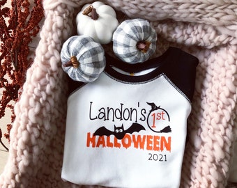 Puede incluir: Camiseta de estilo béisbol blanca y negra con un murciélago negro y el texto "Landon's 1st HALLOWEEN 2021" en la parte delantera.