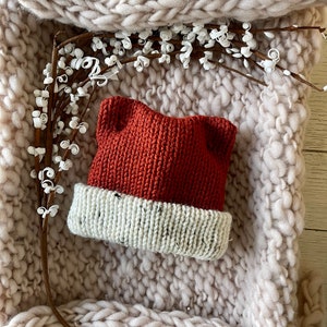 May include: A knitted baby hat with a rust colored top and a white and black speckled brim. The hat is sitting on a cream colored knitted blanket.