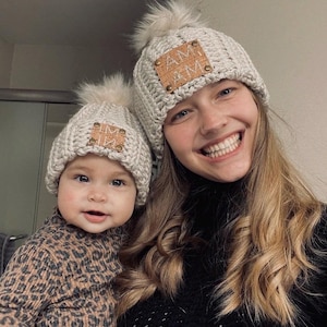 May include: Two matching light gray knit hats with faux fur pom-poms. Each hat has a rectangular leather patch with the initials "AM" and "M" on it. One hat is on a baby, the other on an adult.