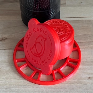 May include: Bright red OXO Rapid Travel Cap set, featuring a bird logo and the text "OXO RAPID TRAVEL CAP". The set includes a lid, a base, and a container, all in a vibrant red color. The base has a circular design.