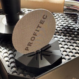 Op de afbeelding: Een accessoire voor een koffiemachine met een cirkelvormig gaasfilter met de tekst "PROFITEC". Het filter wordt vastgehouden door een zwarte, geometrische standaard. De koffiemachine heeft een zwarte buitenkant en een zilveren knop.