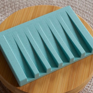 May include: A light blue soap dish with a textured surface and multiple ridges. The dish is made of silicone and is sitting on a wooden base.
