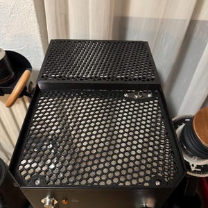 Könnte beinhalten: Schwarze Espressomaschine mit Wabennetzfront und -oberseite. Die Maschine hat silberne Knöpfe und Tasten mit Kontrollleuchten. Der Markenname ist auf der Vorderseite sichtbar. Die Maschine ist für die Zubereitung von Espresso konzipiert.