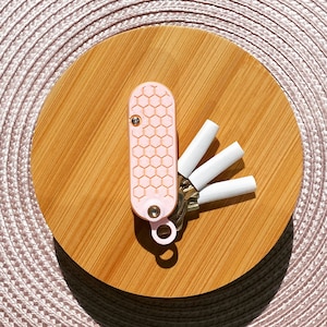 Könnte beinhalten: Ein rosa Schlüsselanhänger mit einem Wabenmuster und einem Satz von vier weißen Schlüsselkappen. Der Schlüsselanhänger befindet sich auf einer Holzoberfläche.