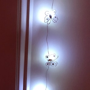 May include: A string of white fairy lights with butterfly-shaped bulbs. The lights are strung along a white wall.