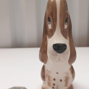 Könnte beinhalten: Eine Keramikfigur eines Basset Hound-Hundes. Der Hund hat braune und weiße Markierungen, eine schwarze Nase und ausdrucksstarke Augen. Die Figur steht aufrecht, mit einer Münze daneben zum Größenvergleich. Die Ohren des Hundes sind lang und schlaff.