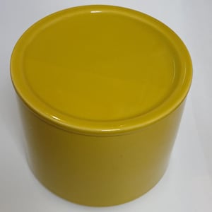 May include: A mustard-yellow ceramic container with a lid. The container is cylindrical with a smooth, glossy finish. The lid fits neatly on top, creating a simple, minimalist design. The container is likely for storage.