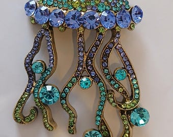 May include: A jeweled jellyfish brooch with a gold-tone metal base. The jellyfish's dome is covered in small, sparkling blue, green, and purple crystals. Large, round blue crystals line the edge, and flowing tentacles feature turquoise and green crystals.