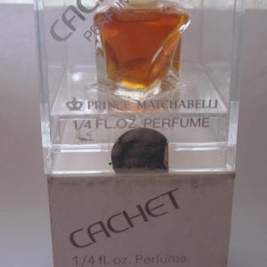 May include: A clear glass bottle of Cachet perfume by Prince Matchabelli. The bottle is in a clear plastic box with a white label that reads "1/4 fl. oz. Perfume" and "0402 00 00".