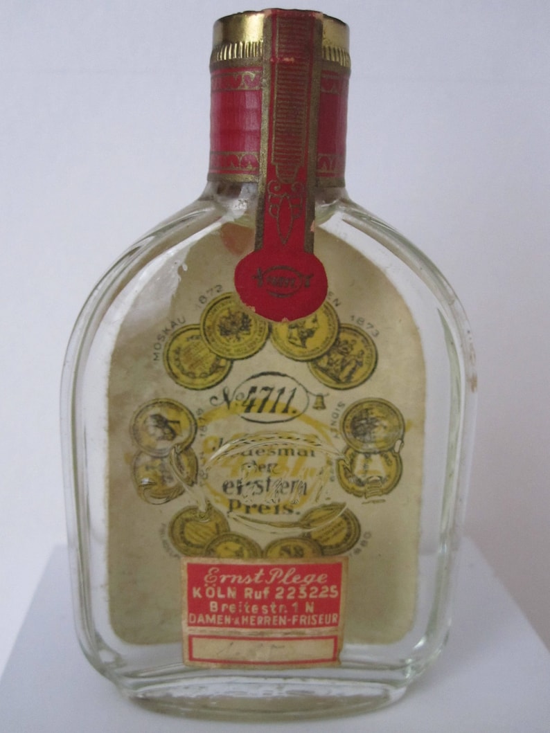 May include: A clear glass bottle with a red and gold label. The label has the text "4711" and "desmai der erster Preis."  The bottle also has a smaller label with the text "Ernst Plege KOLN Ruf 223225 Breitestr. 1N DAMEN HERREN-FRISEUR".