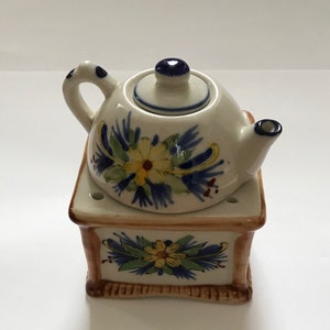 May include: A white ceramic teapot with blue and yellow floral designs sits on a matching base. The teapot has a lid and a spout. The base has a square shape and is decorated with a similar floral design.