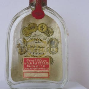 May include: A clear glass bottle with a red and gold label. The label has the text "Jede stal den ersten Droic" and "Ernst Plege K&Ouml;LN Ruf 223225 Breitestr. 1 N DAMEN HERREN-FRISEUR".