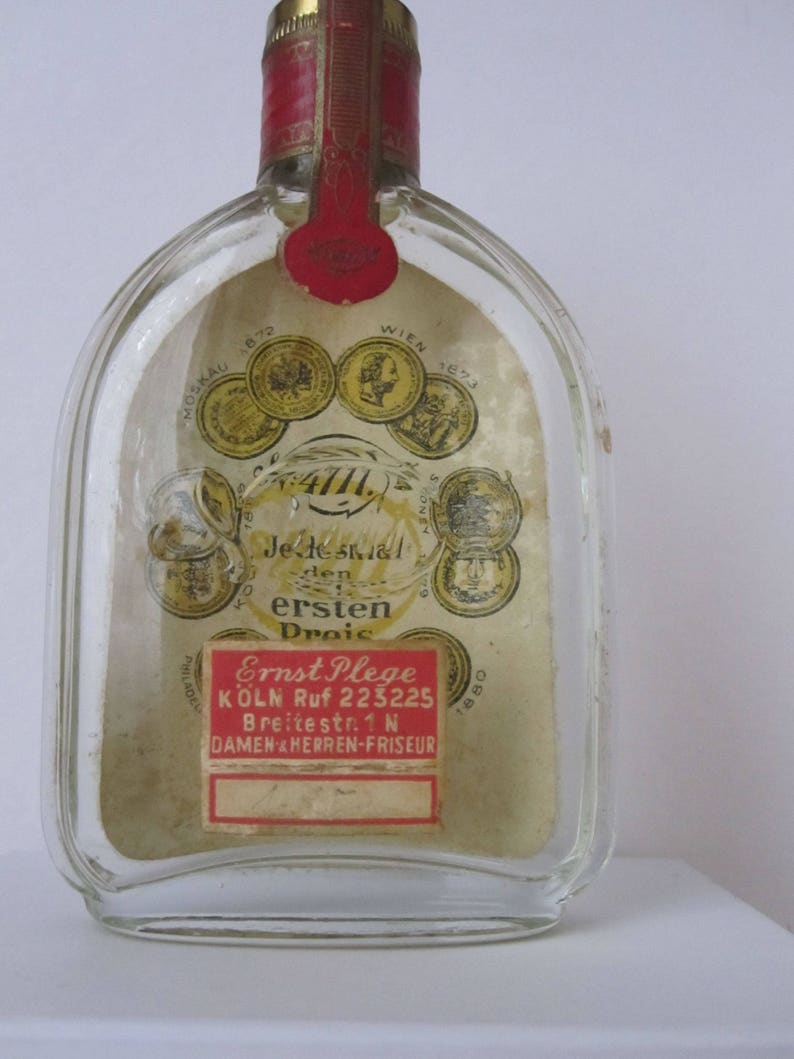 May include: A clear glass bottle with a red and gold label. The label has the text "Jede stal den ersten Droic" and "Ernst Plege K&Ouml;LN Ruf 223225 Breitestr. 1 N DAMEN HERREN-FRISEUR".