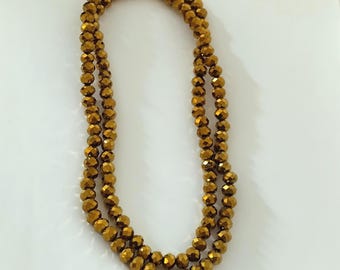 May include: A long strand of metallic gold-colored beads arranged in a double loop. Each individual bead features a faceted, multi-sided surface that reflects light, creating a shimmering appearance throughout the entire length of the necklace.