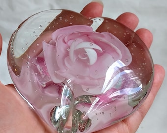 May include: A heart-shaped clear glass paperweight featuring an encased light pink rose with layered petals and a green stem. The glass body contains numerous small air bubbles distributed throughout the transparent material, creating a textured effect.