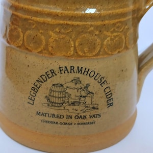 May include: A tan ceramic mug with a handle, featuring the text "LEGBENDER FARMHOUSE CIDER" and an illustration of a barrel and a person. The mug has a decorative apple design around the top and is marked "MATURED IN OAK VATS".