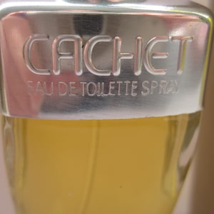 May include: A silver bottle with a silver cap and a yellow liquid inside. The cap has the word "CACHET" embossed on it, along with the words "Eau de Toilette Spray".