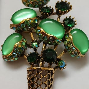 May include: A vintage brooch in the shape of a flower arrangement. The brooch features green cabochon stones and emerald-colored rhinestones set in a gold-tone metal. The flower arrangement is in a gold-tone basket.