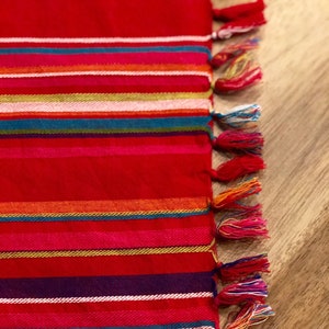 Könnte beinhalten: Roter gestreifter Tischläufer mit bunten Fransen. Der Läufer hat eine Vielzahl von Farben, darunter Rot, Orange, Gelb, Grün, Blau, Lila und Weiß.