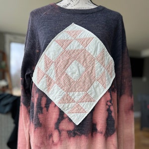Puede incluir: Una sudadera de color gris y rosa con efecto tie-dye y un diseño de patchwork blanco y rosa en la parte delantera. El diseño de patchwork tiene forma de diamante.