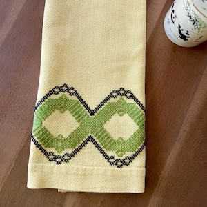 Könnte beinhalten: Ein hellgelbes, rechteckiges Küchentuch aus strukturiertem Waffelpiqué-Gewebe. Der untere Bereich ist mit einem Kreuzstichmuster verziert, das aus zwei ineinandergreifenden Rauten in Grüntönen besteht und mit schwarzem Garn umrandet ist.