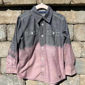 May include: A long-sleeved button-down shirt with a black to pink ombre design. The shirt has two chest pockets and a small embroidered logo on the bottom right.