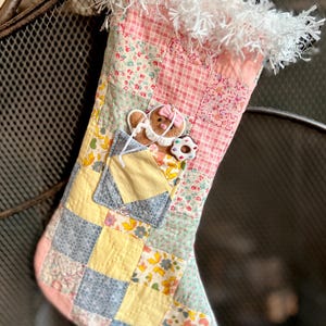 May include: A handmade Christmas stocking with a patchwork design in pastel colors. The stocking features a pocket with a gingerbread person and a donut. The top is trimmed with white fringe.