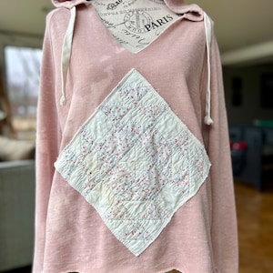 Puede incluir: Una sudadera con capucha rosa con un parche en forma de diamante cosido en la parte delantera. El parche está hecho de tela blanca con un patrón floral rojo y azul.