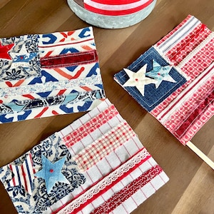 Puede incluir: Tres manualidades textiles rectangulares con diseños de banderas estadounidenses, hechas de tiras superpuestas de telas estampadas en rojo, blanco y azul, encaje y mezclilla. Cada pieza incluye aplicaciones de estrellas de tela y bordes deshilachados.