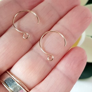 May include: Two gold-colored earring hooks are displayed against a soft, blurred background. The hooks are open at the bottom and have a looped design at the base. The hooks are held in a person's hand.