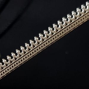 May include: A silver-coloured decorative trim with a repeating crown design. The trim features small, evenly spaced crowns along the top edge and a row of small circular holes along the bottom edge. The background is a solid black.