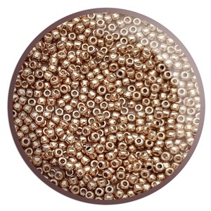 May include: A close-up shot of numerous small, metallic gold seed beads. The beads are tightly packed, filling the circular frame. They have an irregular shape and a shiny surface, reflecting light. Suitable for jewellery making and crafts.