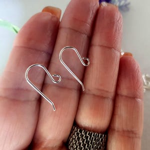 Könnte beinhalten: Zwei silberfarbene Ohrhaken werden vor einem unscharfen Hintergrund gezeigt. Die Haken haben eine gebogene Oberseite und eine gerade, spitz zulaufende Unterseite. Die Haken werden in einer Hand gehalten.