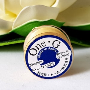 May include: A spool of gold-colored nylon thread labeled "One G" with the text "Made in Japan", "330dtex", "Nylon 100%", "50 yards (約46m)", and "発売元: トーホー株式会社".
