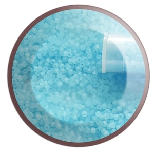 May include: A close-up image of a container filled with light blue seed beads. The beads are small and round, and they are arranged in a random pattern. The container is made of clear plastic and has a white lid.