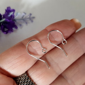 May include: Two silver earring hooks, also known as ear wires, are shown on a hand. The hooks are made of metal and have a simple design.