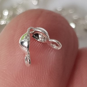 May include: Close-up of a small, silver-coloured jewellery clasp. The clasp is open, revealing a hollow interior. It has a loop on each end for attaching to jewellery components. The clasp is held between two fingers.