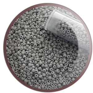 May include: A close-up of a clear plastic tube filled with small, light grey seed beads. The beads are scattered around the tube, creating a textured surface.