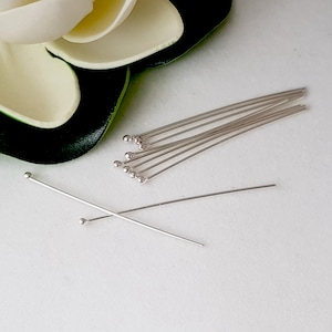 May include: A set of silver head pins with round heads. The pins are arranged in a fan shape on a white surface.