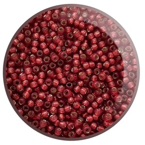 May include: A close-up image of a large quantity of small, round, red seed beads. The beads have a metallic sheen and are arranged in a random pattern.