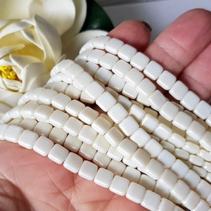 Könnte beinhalten: Eine Strähne weißer quadratischer Perlen. Die Perlen sind aus einem glatten, undurchsichtigen Material gefertigt und haben eine leicht irisierende Oberfläche.