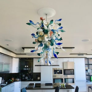 May include: A modern kitchen with white cabinets and a large glass chandelier with blue, green, and white glass pieces. The chandelier is hanging from the ceiling and is centered in the room. The kitchen has a black countertop and stainless steel appliances.