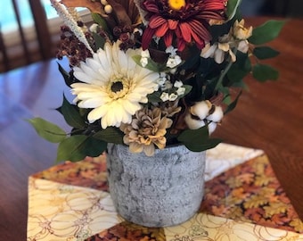 May include: A floral arrangement in a textured gray pot featuring a large dark red daisy, a white daisy, cotton bolls, and dried foliage. The arrangement sits on a hexagonal fabric mat with a pattern of pumpkins and autumn leaves. Text reads Fall Leaves.