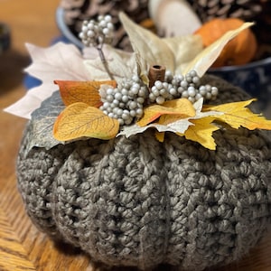 May include: A crocheted olive green pumpkin featuring a textured yarn surface. The top is decorated with artificial autumn foliage, including yellow and orange leaves, clusters of small white berries, and a small brown cinnamon stick stem.
