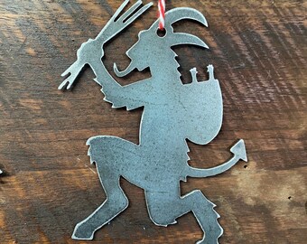 May include: A metal ornament shaped like a devil holding a pitchfork and a sack. The ornament is painted silver and has a red and white string attached to it.