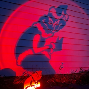 May include: A red-lit silhouette of a character is projected onto a white wall. The character is holding a small object, with a matching illuminated metal cutout at the base. The scene is set outdoors at dusk.