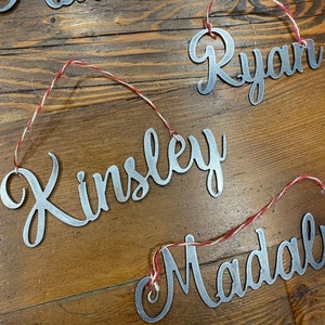 Puede incluir: Un banner de metal con los nombres Matthew, Ryan, Kinsley y Madal escritos en letra cursiva. El banner está encordado con hilo rojo y blanco.