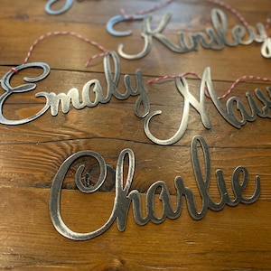 Puede incluir: Cinco letreros de metal con nombres en letra cursiva, colgados de una cuerda roja y blanca. Los nombres son Justin, Kinsley, Emily, Max y Charlie.